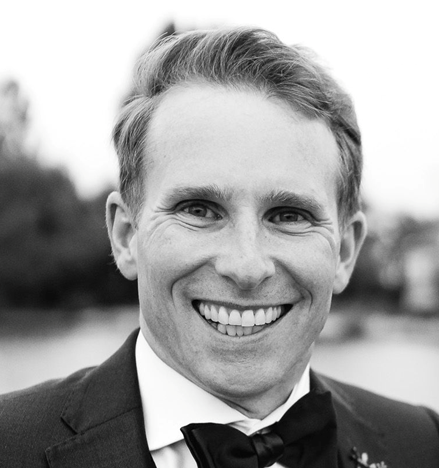 fotografía en primer plano, en blanco y negro, de un hombre que sonríe ante cámara con un traje de chaqueta